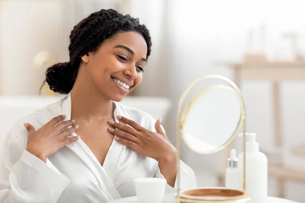 Beautiful black woman looking in mirror and touching decollete zone, smiling african american female in white silk robe checking wrinkles on neck, enjoying domestic beauty treatments, copy space