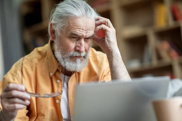 Pensive grey-haired bearded old man in casual outfit using modern notebook, reading article, surfing on Internet, touching his head, holding eyeglasses, home interior, closeup, copy space