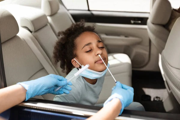 Black Preteen Girl Getting Tested For Covid-19 Sitting In Car, Traveling With Family By Automobile. Unrecognizable Medical Worker Making PCR Test For Child At Drive-Thru Diagnostic Center.