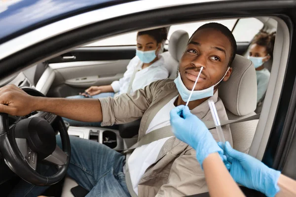 African American Man Getting Tested For Coronavirus In Car, Driving Traveling With Family By Auto. Unrecognizable Medical Worker Making PCR Test For Male Driver. Covid-19 Pandemic Concept