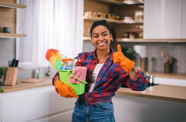 身穿橡胶手套 头戴一桶清洁用品的快乐的年轻非洲裔美国女人露出大拇指 认可在现代厨房内部进行清洁 卫生和家庭主妇应享有的福利 — 图库照片