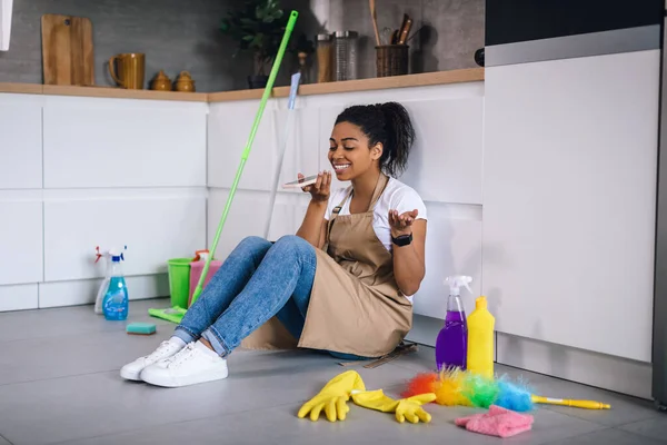 Smiling young african american lady sit on floor with cleaning supplies and talking on phone at modern kitchen interior. Cleaning, housework, hygiene and housewife at home, call and gossip with friend