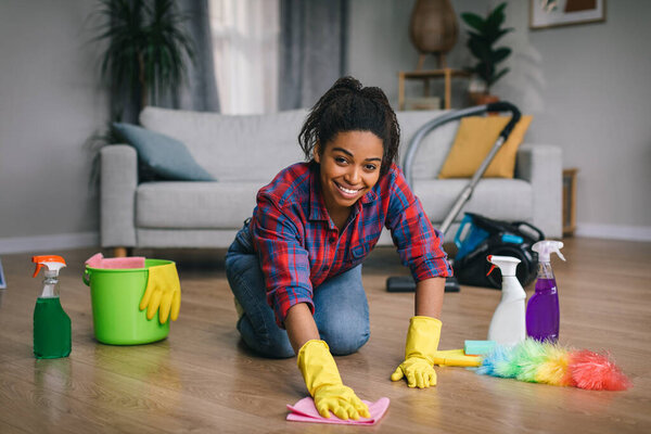 Cheerful young african american lady in rubber gloves with cleaning supplies washes floor at modern room interior, free space. Cleaning alone, housework, hygiene and housewife at home due covid-19
