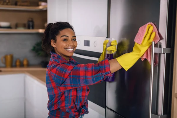 ゴム手袋の幸せな若いアフリカ系アメリカ人女性は 現代のキッチンのインテリアでスプレーとスポンジで冷蔵庫を拭く 主婦の仕事での清掃 Covid 19パンデミック — ストック写真