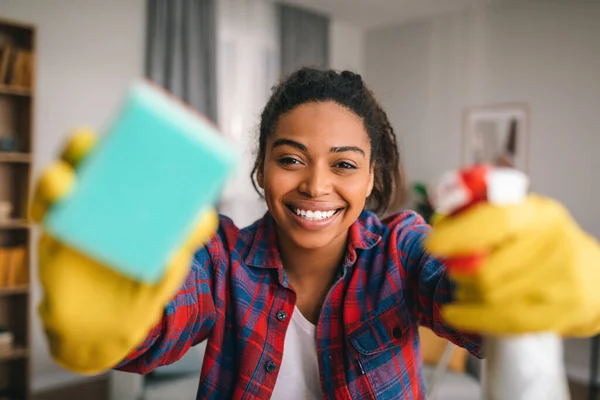 Smiling young african american lady in rubber gloves with spray and sponge wipes camera at modern kitchen interior. Perfectionism, cleaning, housework, hygiene and housewife have fun at home, covid-19