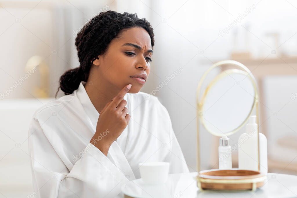 Frustrated Black Woman With Problem Skin Looking At Pimple On Cheen While Sitting In Front Of Mirror In Bathroom, Confused African American Female In White Silk Robe Suffering Acne, Closeup