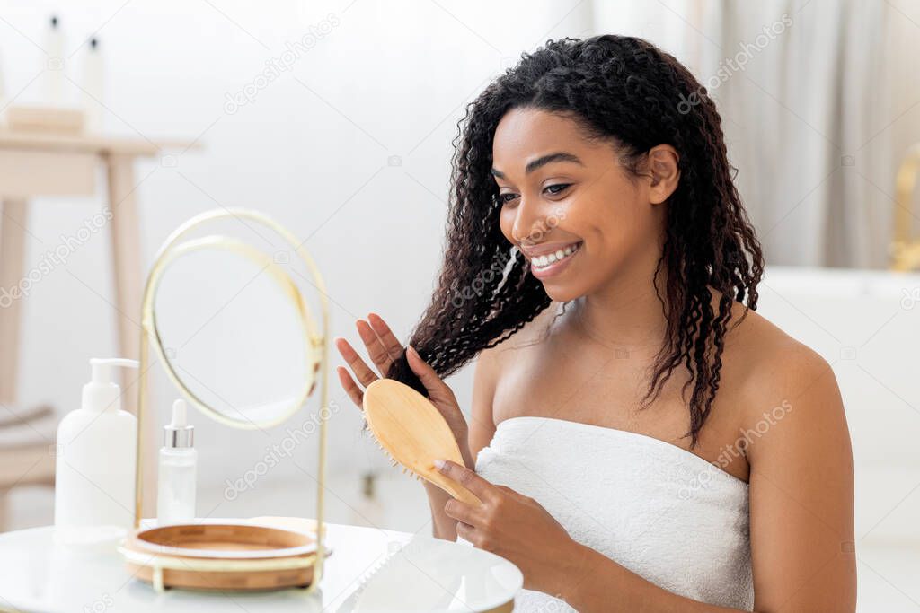 Smiling Young Black Woman Brushing Her Beautiful Curly Hair With Comb While Sitting Near Mirror In Bathroom, Happy African American Female Wrapped In Towel Making Haircare Routine At Home