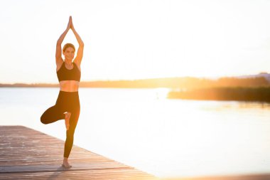 Sporty middle aged female practicing yoga on a wooden pier on sunset, slim beautiful woman meditating outdoors near lake, smiling lady standing in tree pose, enjoying wellness and healthy lifestyle