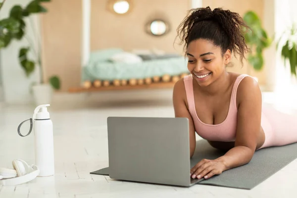 Happy young black lady lying on mat and using laptop computer, talking to trainer on web or watching sports video, working out at home, copy space