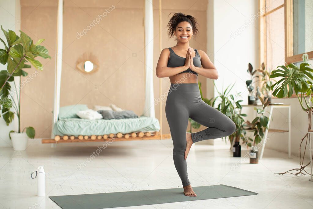 Life balance. Happy black lady practicing yoga, doing tree pose, working out on fitness mat at home, copy space. Positive woman in activewear standing on one leg with hands clasped