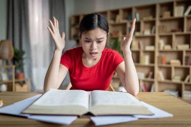 Big mistake. Amazed unhappy teenager chinese girl reading book, gesticulate in room or library interior. Human emotions, shocked facial expression, prepare for exam at home during covid-19 outbreak