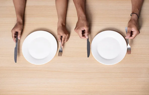 Hands of hungry millennial african american woman and man hold cutlery near empty plates on table in kitchen. Waiting for dinner, problems with eat, crisis, no food and starving