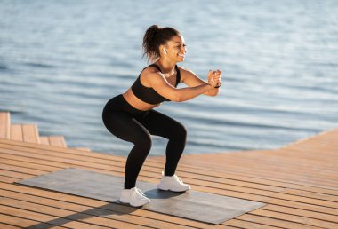 Geniş yapılı, dışarıda antrenman yapan, Afro-Amerikan spor elbiseli, siyah spor ayakkabılı, beyaz spor ayakkabılı, gölün yanındaki parkta yoga minderi üzerinde egzersiz yapan, fotokopi çeken bir kadın.