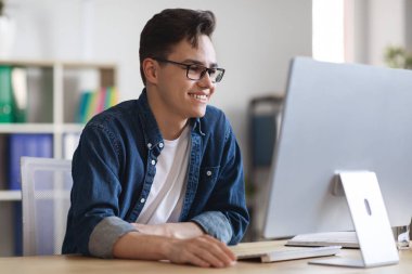Portrait Of Smiling Millennial Businessman Working On Computer In Office, Happy Young Male Entrepreneur Sitting At Desk And Looking At Monitor, Browsing Internet At Workplace, Closeup Shot