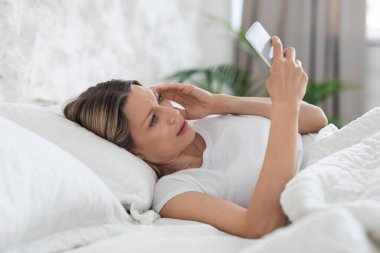 Unhappy blonde young woman lying alone in bed at home, using modern smartphone and touching her head, surfing on Internet, seeking for treatment medicine against headache, side view