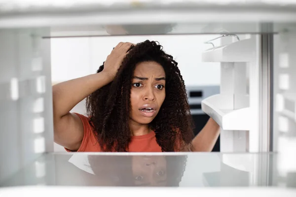 Hungry shocked disappointed pretty millennial african american woman with open mouth checks empty fridge in kitchen, close up. Facial expression, crisis, human emotions and no food at home, free space