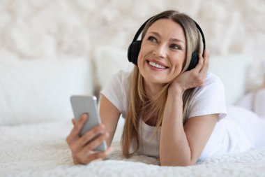 Evdeki modern akıllı telefon ve kablosuz müzik kulaklığı kullanan hayalperest sarışın genç bayan. rahat beyaz yatak odasında uzanmış fotokopi odasına bakıyor ve gülümsüyor, eğlenceli uygulamaları kontrol ediyor, kapatıyor.