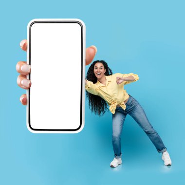 Mobile Offer. Positive Arabic Lady Showing Smartphone Empty Screen To Camera Advertising Application Posing In Studio Over Blue Background. Mockup, Square Shot