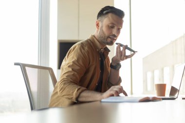 İş yerinde erkek girişimcinin ses arama uygulamasını kullanışının yan görüntüsü. Ofiste çalışan bir iş adamı yüksek sesle konuşma modunda. Modern İletişim Konsepti