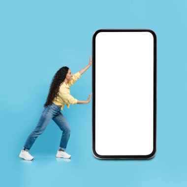 Middle Eastern Lady Posing With Huge Cellphone Blank Screen Advertising Mobile Offer Over Blue Studio Background. Cheerful Woman Pushing Large Telephone. Mockup, Square Shot