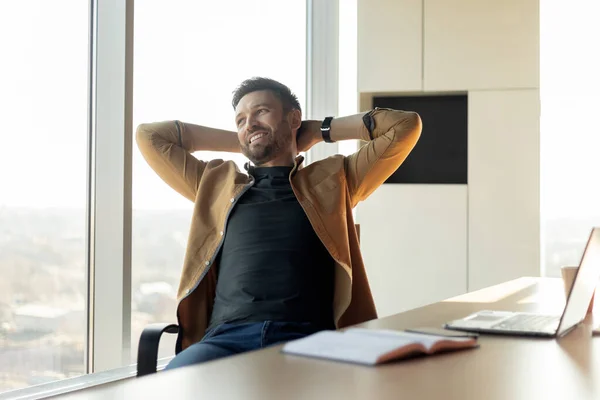 Successful Business Career. Happy Male Entrepreneur Relaxing In Chair Holding Hands Behind Back Sitting In Modern Office Indoor, Looking Aside. Businessman Resting After Work At Workplace