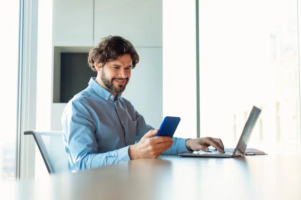 Cheerful mature businessman checking smartphone, using laptop computer, working online at company office, blank space. Professional occupation, business lifestyle concept