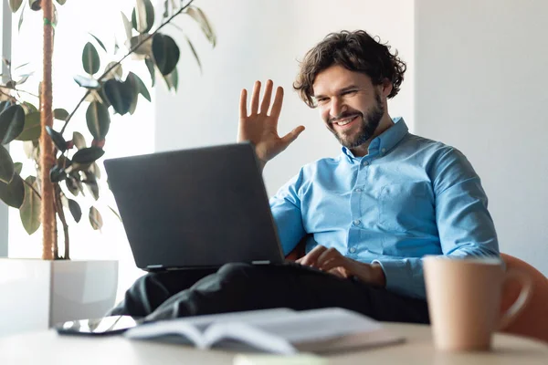 Uzaktan iletişim, sanal buluşma konsepti. Resmi kıyafetleri içinde mutlu bir iş adamı dizüstü bilgisayarda video çekiyor, kameraya el sallıyor ve gülümsüyor, modern ofiste oturuyor.
