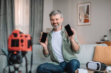 Happy caucasian old man with beard shows smartphones with blank screens and makes video review in living room interior. Technology comparison, hobby at home, modern blog during covid-19 pandemic