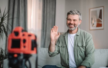 Smiling caucasian mature male with beard waving hand at camera, shooting video in living room interior. Modern work remote, new tech, hobbies and new normal, blogging at home due covid-19 quarantine