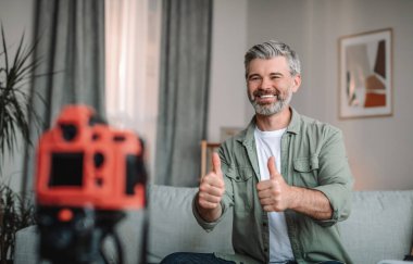 Glad happy caucasian old male blogger with beard showing thumbs up and shooting video in living room interior. New technology, hobbies and new normal, blog and social distance due covid-19 pandemic