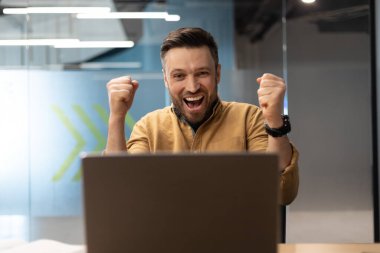 Emotional Business Man Shouting Shaking Fists In Joy Using Laptop Reading Great News Online Smiling To Camera, Celebrating Success At Workplace. Male Office Worker Gesturing Yes.