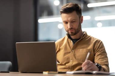 Ciddi bir iş adamı dizüstü bilgisayar kullanıyor, iş raporları yazıyor ve modern ofisteki masada oturan notlar alıyor. Şirket yaşam tarzı, girişimcilik kariyeri.