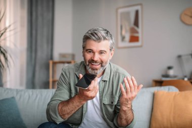 Glad happy emotional caucasian mature male with beard speaks by phone in living room interior. Pensioner calls, communication at home, social distancing and tech, new normal during covid-19 pandemic