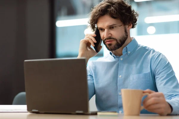 Mature businessman in formal wear talking on smartphone, enjoying conversation at workplace, copy space. Attractive middle aged entrepreneur speaking with customer while working on laptop at office