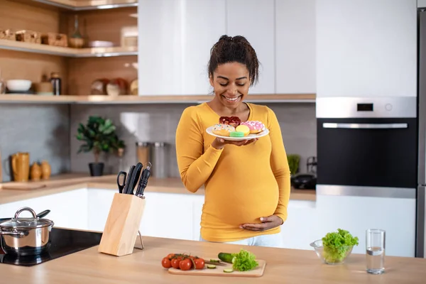 Happy young pregnant black lady touching big belly, hold plate with sweets at table with vegetables and glass of water in kitchen interior. Sweet tooth and desire for junk food, health care and diet