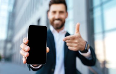 Smiling european young bearded businessman in suit rejoices to success and points finger at smartphone with blank screen near office building. Online victory, recommendation of new app and website