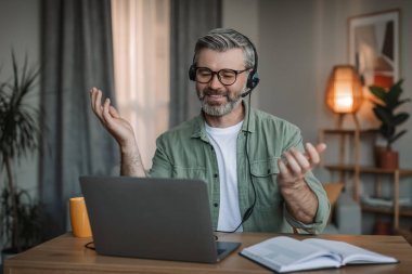 Gözlüklü, sakallı, kulaklıklı, gülümseyen beyaz, yaşlı bir erkek. Dizüstü bilgisayara bakıyor. Oda içindeki müşterileri uzaktan destekliyor. Sosyal mesafe, emekliler evde çalışıyor, toplantı ve iş gereği covid-19