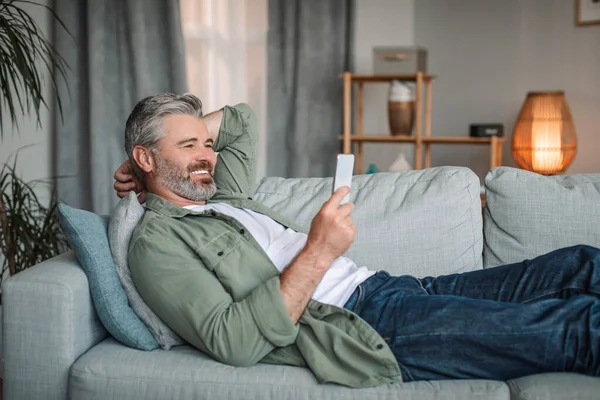 Cheerful caucasian old man with beard watch video on smartphone, rest and relax at sofa alone in living room interior. Video call, new app, online offer and technology at home due covid-19 pandemic