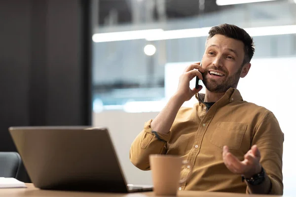 Happy Businessman Speaking By Phone Sitting At Laptop Working Online In Modern Office. Man Communicating Talking On Cellphone At Workplace. Business And Mobile Communication Concept