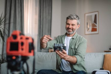 Blogging at home. Happy caucasian senior man with beard shows photo camera and shoots video in room interior. Review on tech, hobbies and new normal, distance work for retired due covid-19 pandemic
