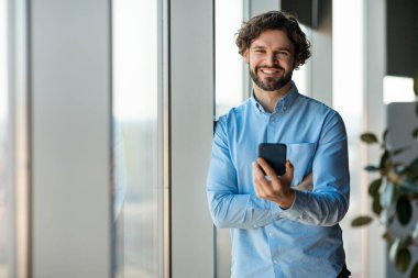 Akıllı telefondan internete girip modern ofisin büyük penceresinin yanında duran mutlu girişimci, kopyalama alanı. Gülümseyen orta yaşlı iş adamı cep telefonuyla poz veriyor. İş iletişimi
