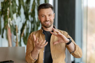 İş İletişimi. Erkek Girişimci Kameraya Bakarken Gülümseyen Bir Şey Açıklıyor Video aracılığıyla Modern Ofiste İşyerinde Oturuyor