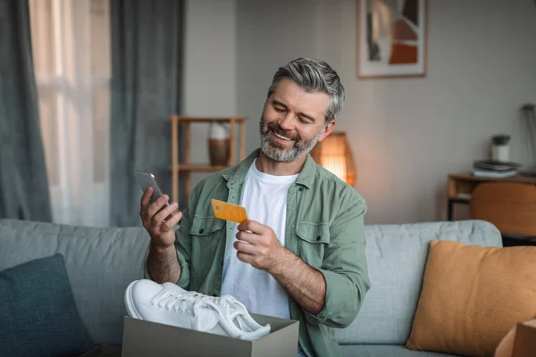 Glad caucasian old man with beard with cardboard box with sneakers uses credit card and smartphone in room interior. Big sale, delivery order, online shopping, cashback at home due covid-19 pandemic