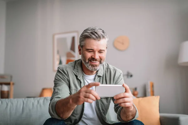 Cheerful caucasian old man with beard watching video on phone in living room interior. Pensioner have video call, new app, advertisement and offer at spare time with device during covid-19 pandemic
