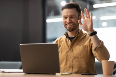 Cheerful Male Entrepreneur Waving Hello To Laptop Making Video Call Sitting At Workplace. Businessman Communicating Online In Modern Office. Business Communication Concept