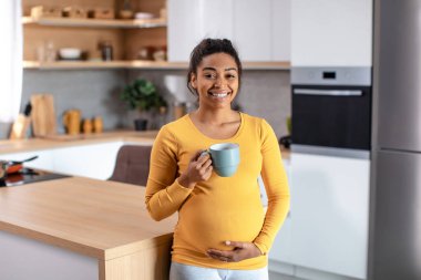 Mutlu, genç, hamile, Afro-Amerikan kadın, koca göbeğine elleriyle dokunuyor, en sevdiği içeceği içiyor ve minimalist mutfağın içinde çayın tadını çıkarıyor. Evde dinlenme ve dinlenme.