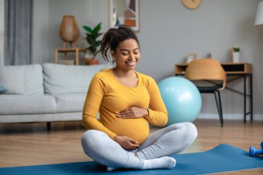 Genç hamile Afrikalı Amerikalı kadının koca göbeğine dokunması ve bebek hareketinin keyfini çıkarması ve oturma odasının içinde yoga yapması güzel. Hamilelik boyunca hassas anlar, konforlu evde ve annelikte egzersiz.