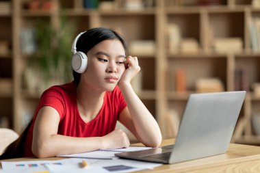 Kablosuz kulaklıklı üzgün Asyalı kız öğrenci oda içinde ders çalışan dizüstü bilgisayara bakıyor. covid-19 salgını sırasında eğitim ve iş sorunları, sosyal uzaklık ve evde aşırı çalışma