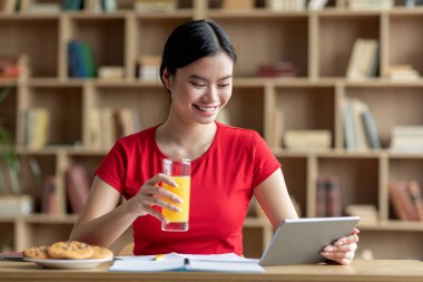 Mutlu Asyalı genç bayan tablette video dersi izliyor, internette sörf yapıyor, meyve suyu içiyor, oda içinde kahvaltı yapıyor. Modern cihaz, atıştırmalık işi, evde çalışma için yeni uygulama, covid-19 salgını