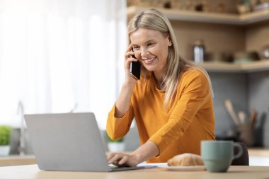 Happy attractive blonde woman entrepreneur talking on cell phone, working on laptop, having conversation with her assistant or client while having breakfast, kitchen interior, copy space clipart
